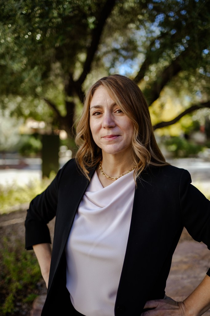 Professor Eve Hanan smiles with her hands on her hips. She has long brown hair and wears a black blazer with a white blouse.